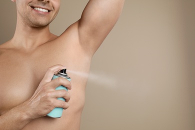 Young man applying spray deodorant to armpit on beige background, closeup. Space for text