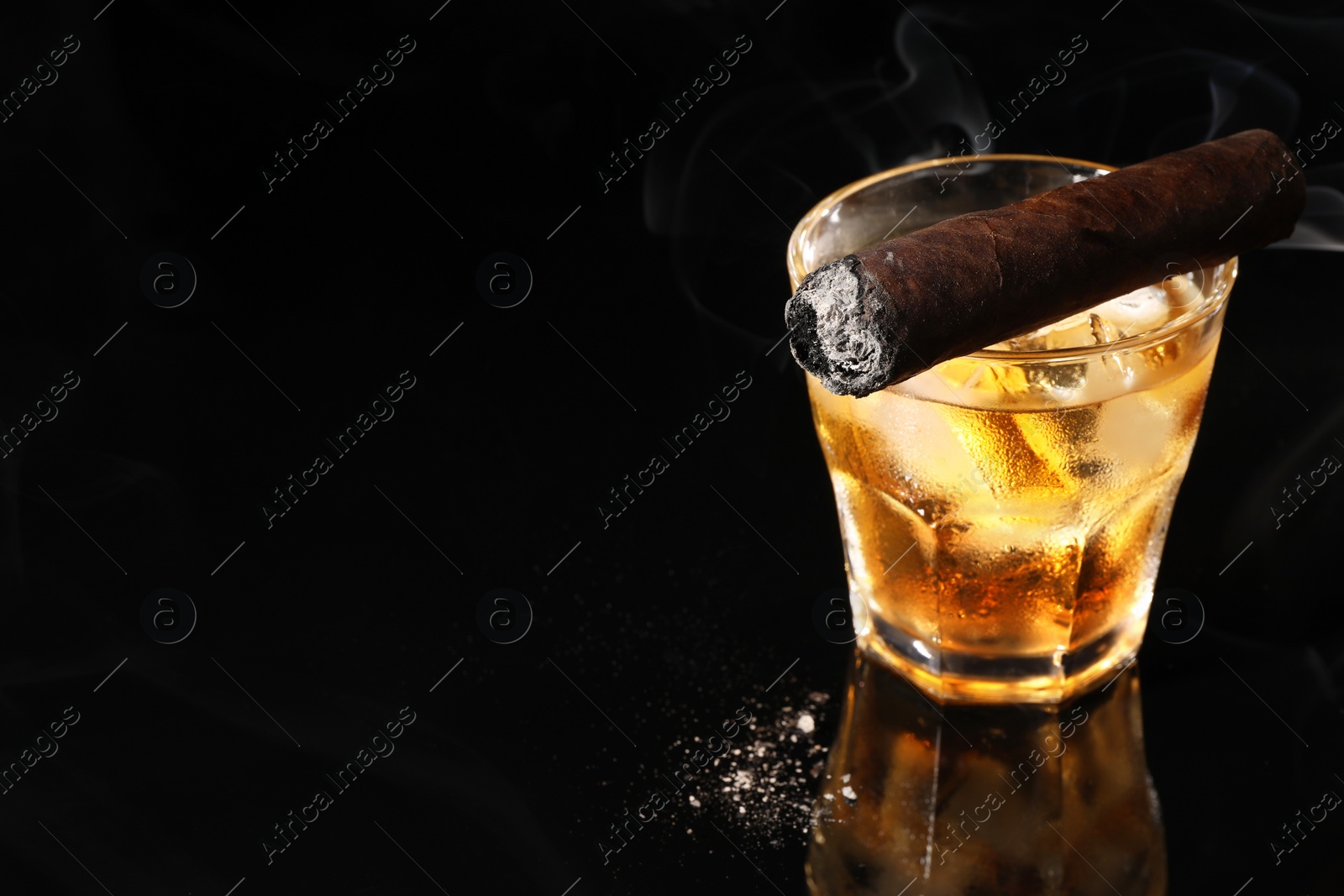 Photo of Glass of whiskey with ice cubes and smoldering cigar on black background. Space for text