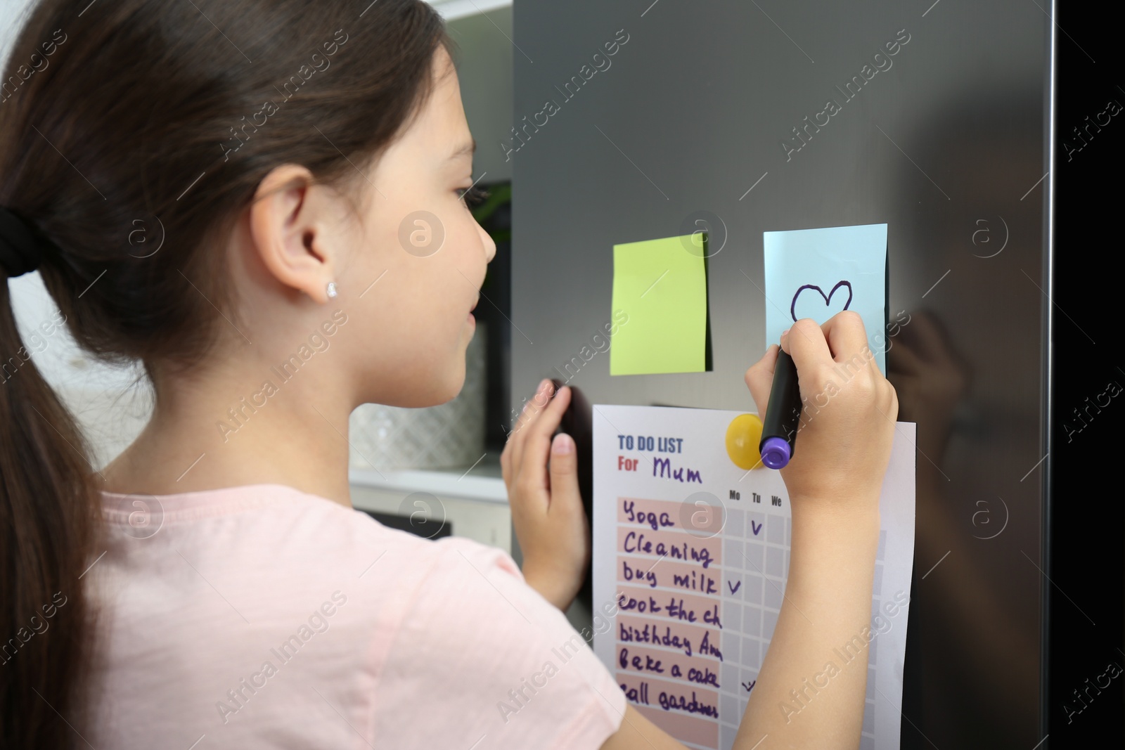 Photo of Little girl drawing heart on note near to do list in kitchen, closeup