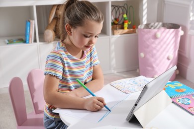 Photo of Little girl learning English with tablet indoors at online lesson