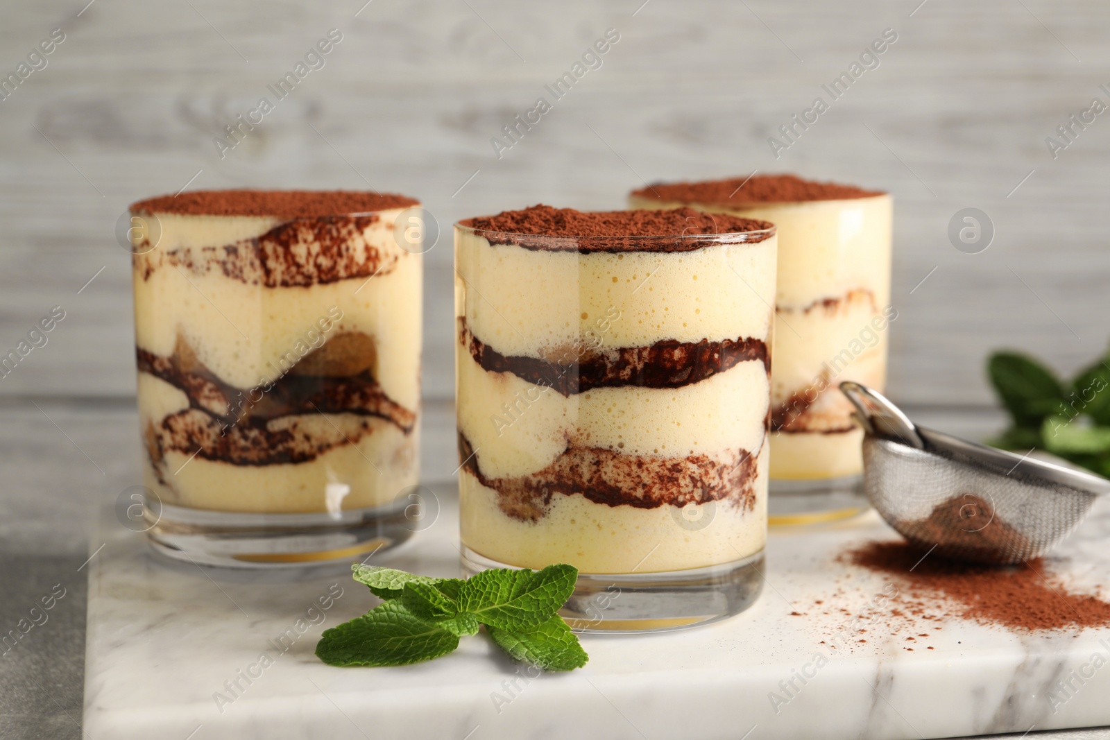 Photo of Delicious tiramisu in glasses, mint and sieve with cocoa powder on table, closeup