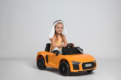Photo of Cute little girl in pilot hat driving children's electric toy car on grey background