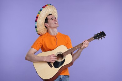 Young man in Mexican sombrero hat playing guitar on violet background