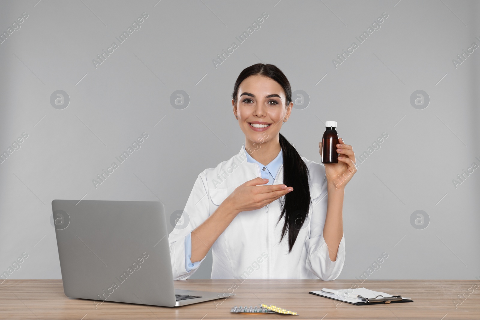 Photo of Professional pharmacist with syrup and laptop at table against light grey background