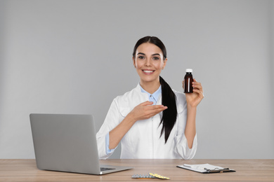 Professional pharmacist with syrup and laptop at table against light grey background