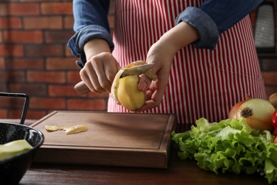 Photo of Woman peeling fresh potato with knife at wooden table indoors, closeup