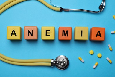 Photo of Word Anemia made with wooden cubes, pills and stethoscope on light blue background, flat lay