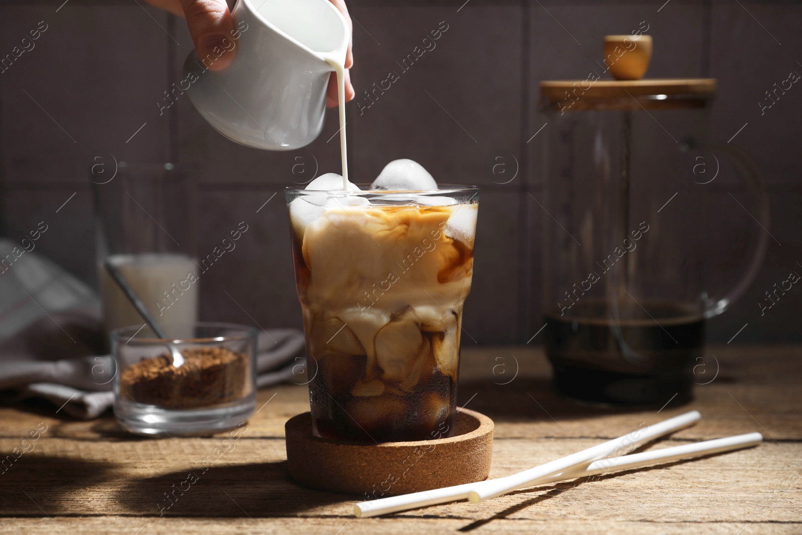 Photo of Woman pouring milk into glass with refreshing iced coffee at wooden table, closeup