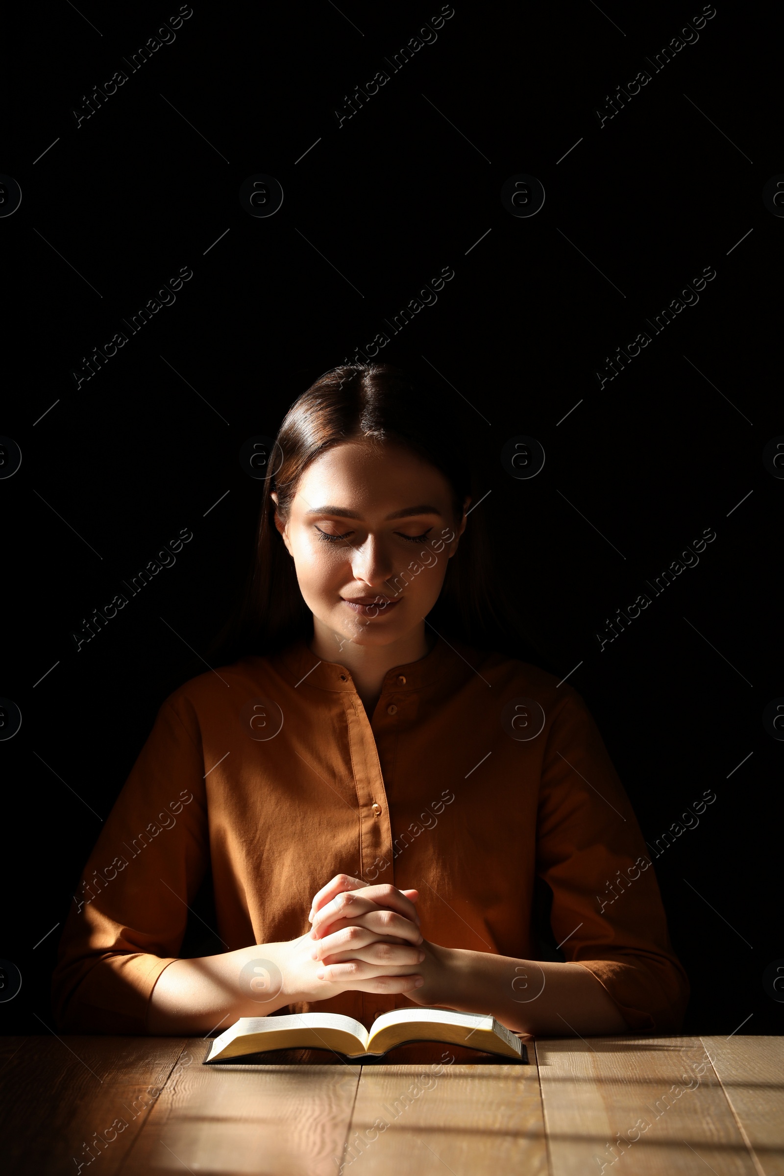 Photo of Religious young woman praying over Bible at wooden table indoors