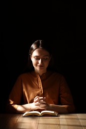 Photo of Religious young woman praying over Bible at wooden table indoors