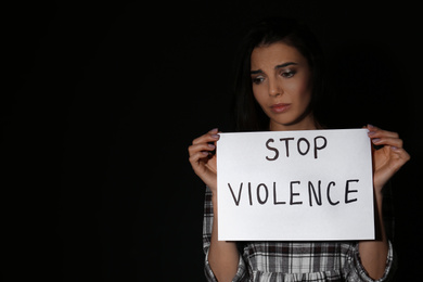 Abused young woman with sign STOP VIOLENCE on black background, space for text