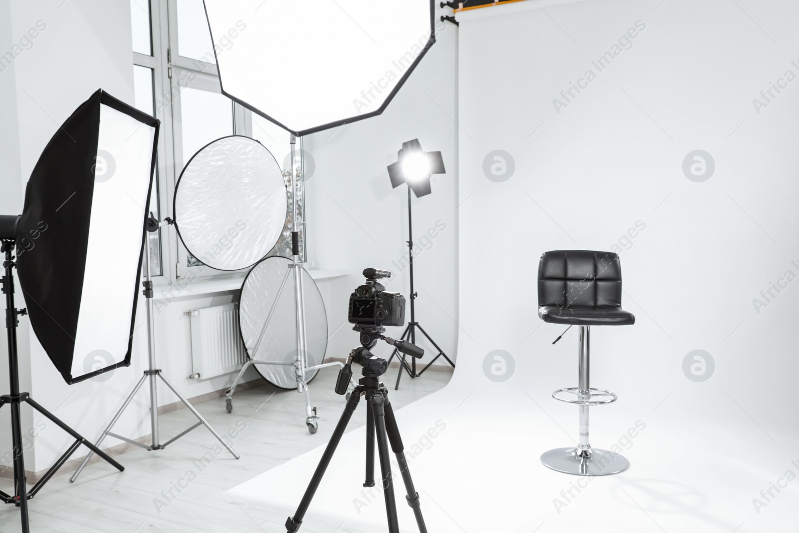 Photo of Tripod with camera, bar stool and professional lighting equipment in modern photo studio