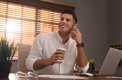 Photo of Freelancer with cup of coffee talking on phone while working at table indoors