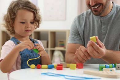 Motor skills development. Father and daughter playing with wooden pieces and string for threading activity at table indoors, closeup