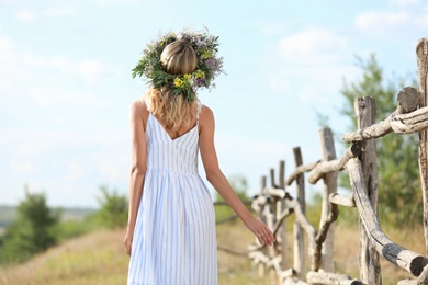 Photo of Young woman wearing wreath made of beautiful flowers near wooden fence on sunny day, back view