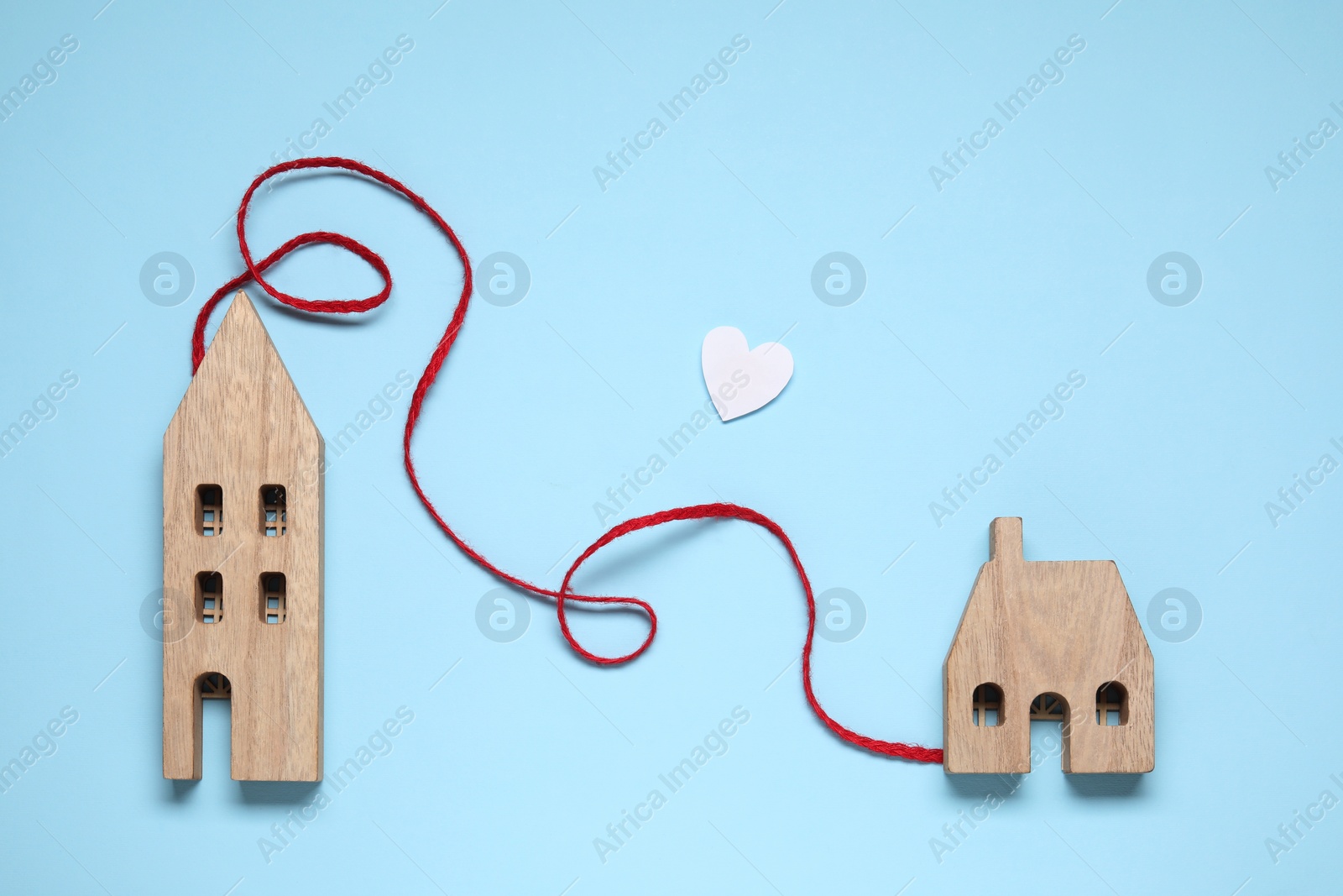 Photo of Red decorative cord and paper heart between two wooden house models on light blue background symbolizing connection in long-distance relationship, flat lay