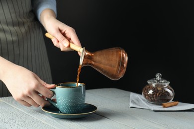 Photo of Turkish coffee. Woman pouring brewed beverage from cezve into cup at gray wooden table against black background, closeup