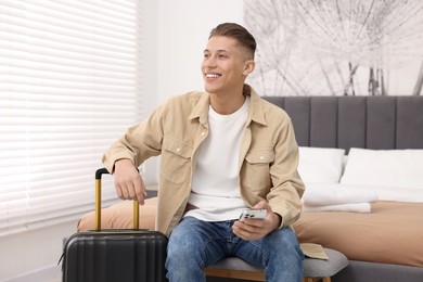 Photo of Smiling guest with smartphone and suitcase in stylish hotel room
