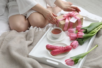 Photo of Tasty breakfast served in bed. Woman with gift box, tea, eclairs, flowers and I Love You card at home, closeup