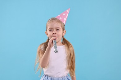 Birthday celebration. Cute little girl in party hat with blower on light blue background