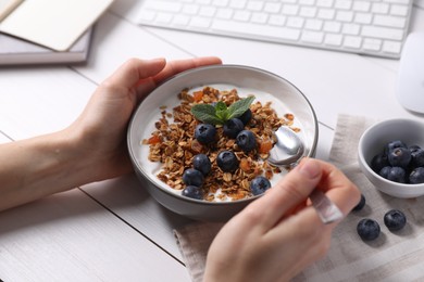 Woman holding bowl of tasty granola with blueberries at white wooden table, closeup