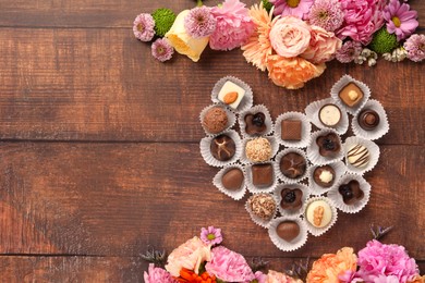 Heart made with delicious chocolate candies and beautiful flowers on wooden table, flat lay. Space for text