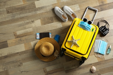 Flat lay composition with suitcase, protective masks, antiseptic spray and personal items on wooden floor. Travelling during coronavirus pandemic