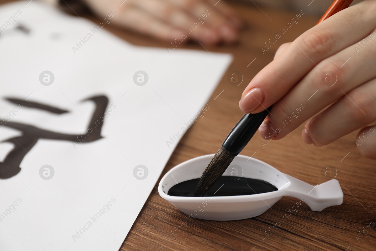 Photo of Calligraphy. Woman dipping brush into inkwell at wooden table, closeup