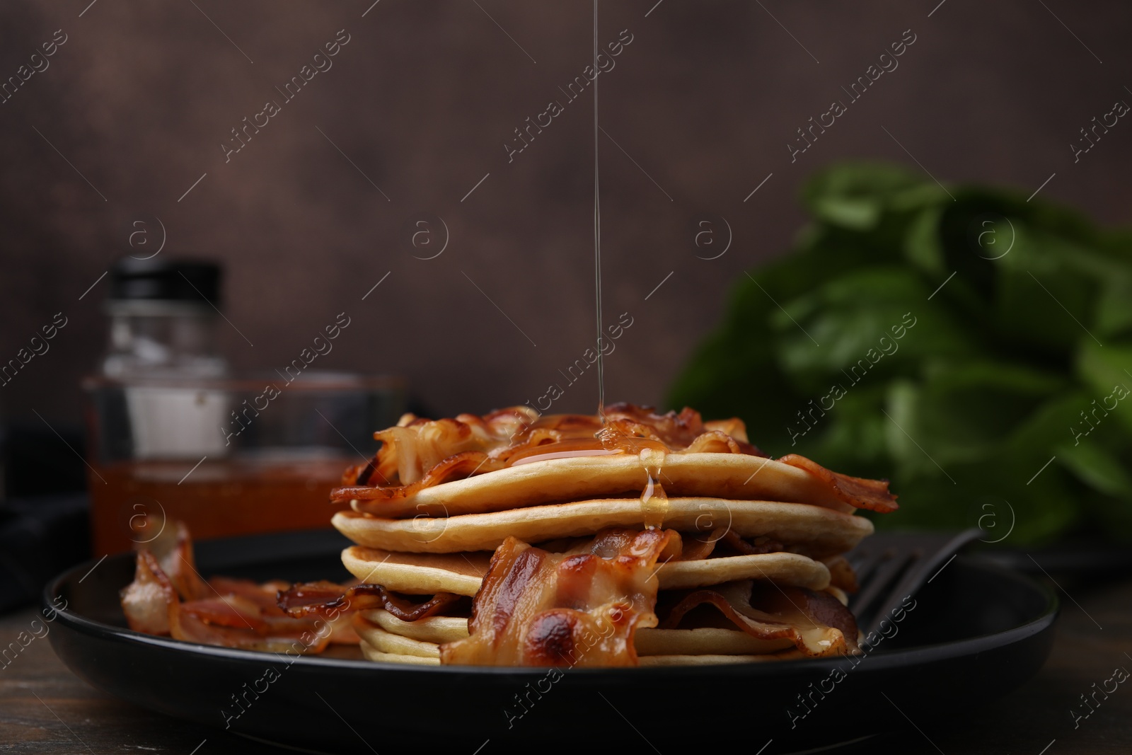 Photo of Pouring maple syrup onto delicious pancakes with fried bacon on wooden table, closeup