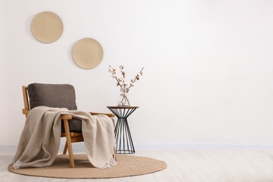 Photo of Comfortable armchair, blanket, side table and vase with cotton branches near white wall indoors. Space for text