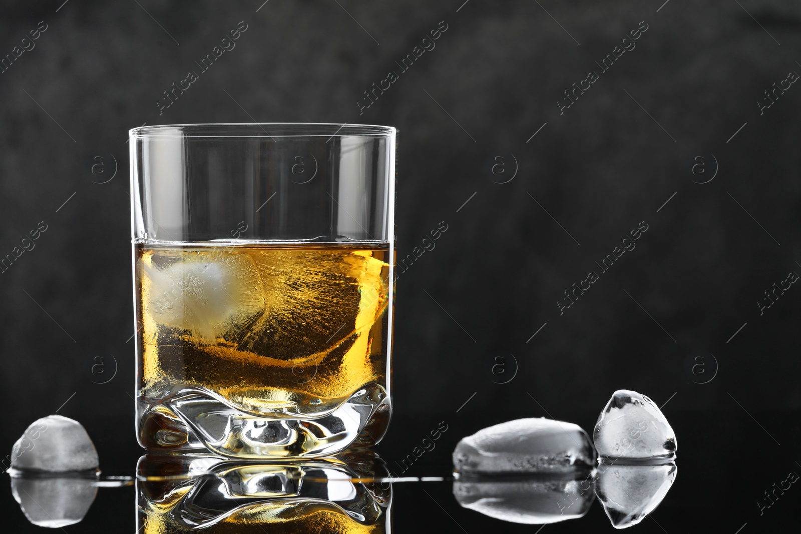 Photo of Tasty whiskey and ice cubes in glass on mirror table against black background, closeup. Space for text