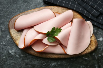 Photo of Board with slices of tasty boiled sausage and parsley on dark textured table, top view