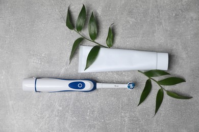 Photo of Electric toothbrush, tube of paste and green leaves on grey textured table, flat lay