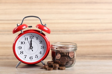 Photo of Red alarm clock, glass jar and coins on table against wooden background. Money savings