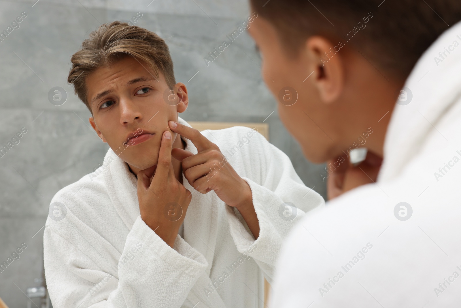 Photo of Upset young man looking at mirror and popping pimple on his face indoors. Acne problem