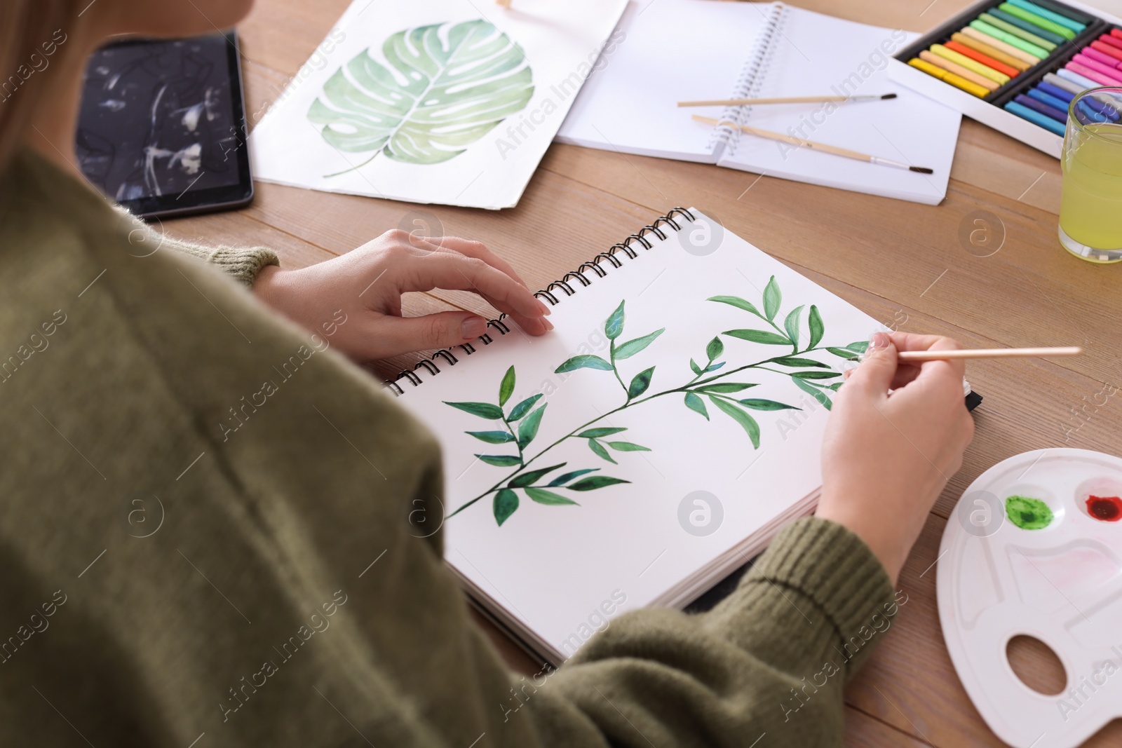 Photo of Woman painting green twig in sketchbook at wooden table, closeup