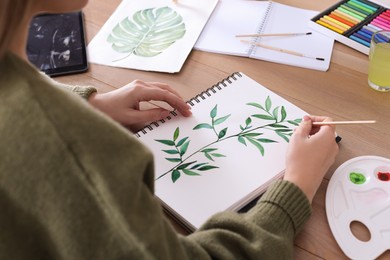 Photo of Woman painting green twig in sketchbook at wooden table, closeup