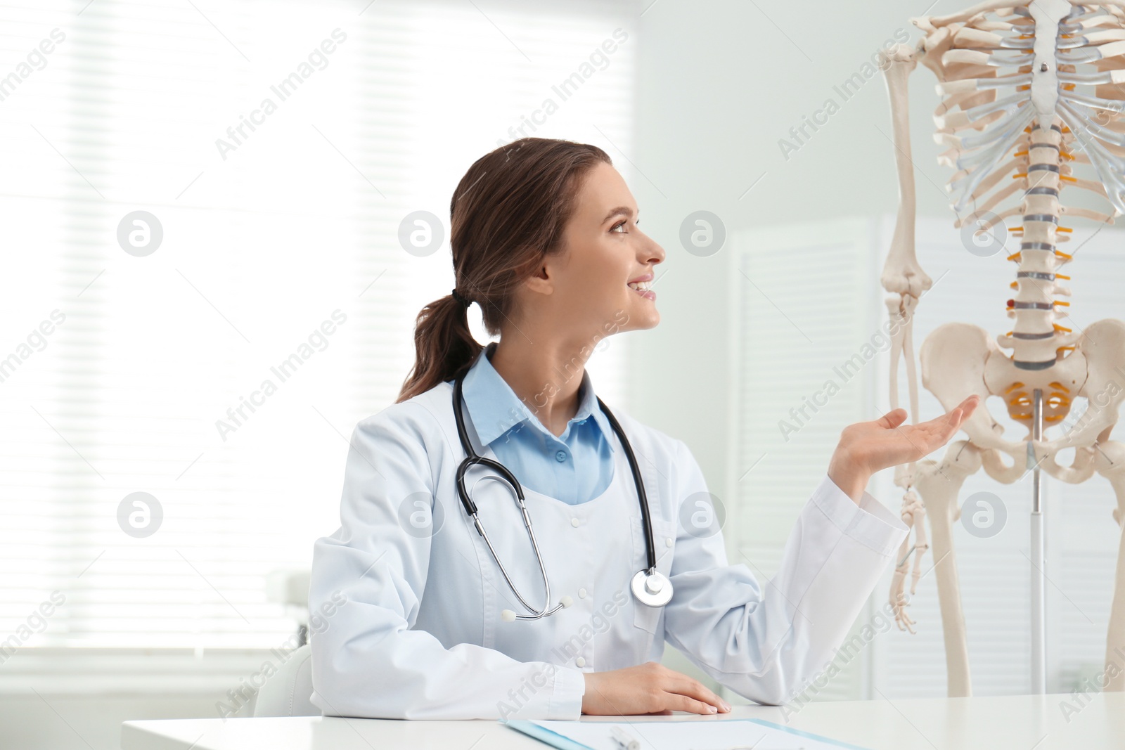 Photo of Female orthopedist at table near human skeleton model in office