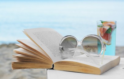 Photo of Open book, sunglasses and glass of refreshing drink on white table near sea