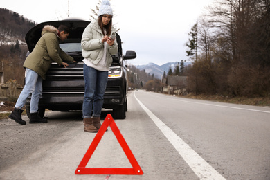 Photo of Stressed couple near broken car and emergency stop sign outdoors. Winter day