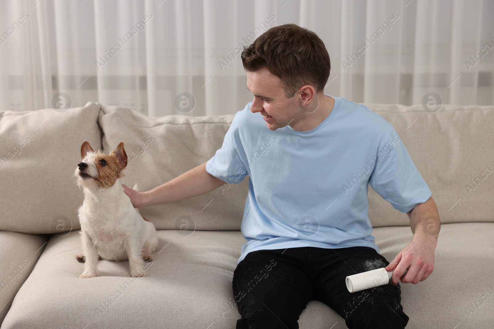 Photo of Pet shedding. Smiling man with lint roller removing dog's hair from pants on sofa at home