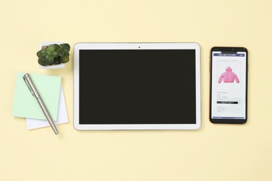 Online store website on device screen. Tablet, smartphone, stationery and houseplant on beige background, flat lay