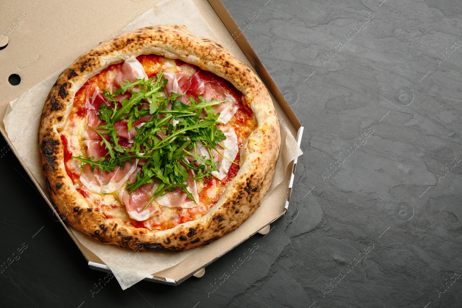 Photo of Tasty pizza with meat and arugula in cardboard box on black table, top view. Space for text