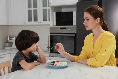Mother feeding her son in kitchen. Little boy refusing to eat dinner