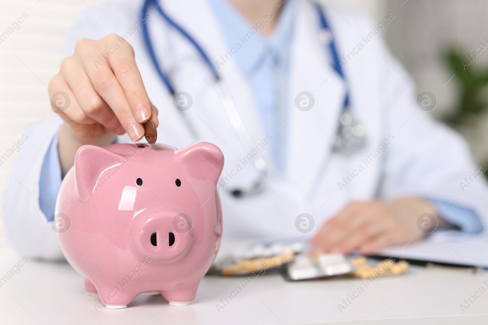 Photo of Doctor putting coin into piggy bank at white table indoors, closeup