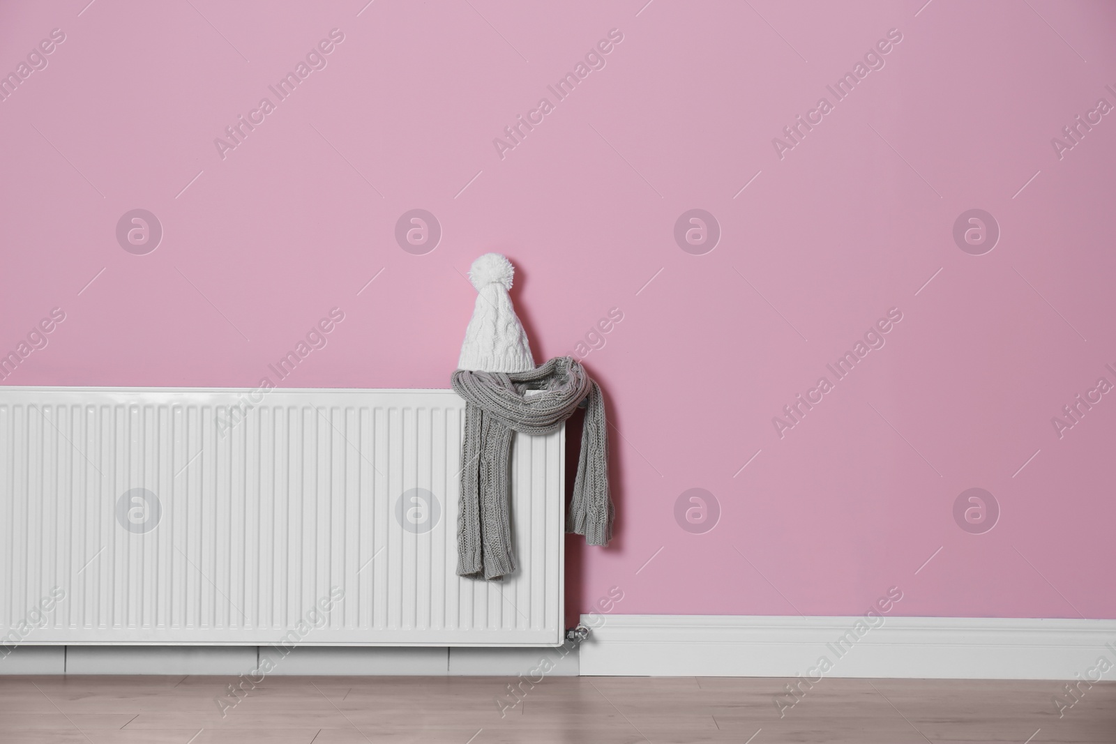 Photo of Knitted hat and scarf on modern radiator near color wall with space for text. Central heating system