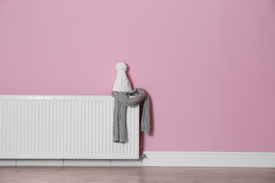 Photo of Knitted hat and scarf on modern radiator near color wall with space for text. Central heating system