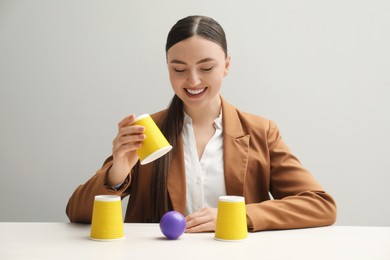Shell game. Happy woman showing ball under cup at white table