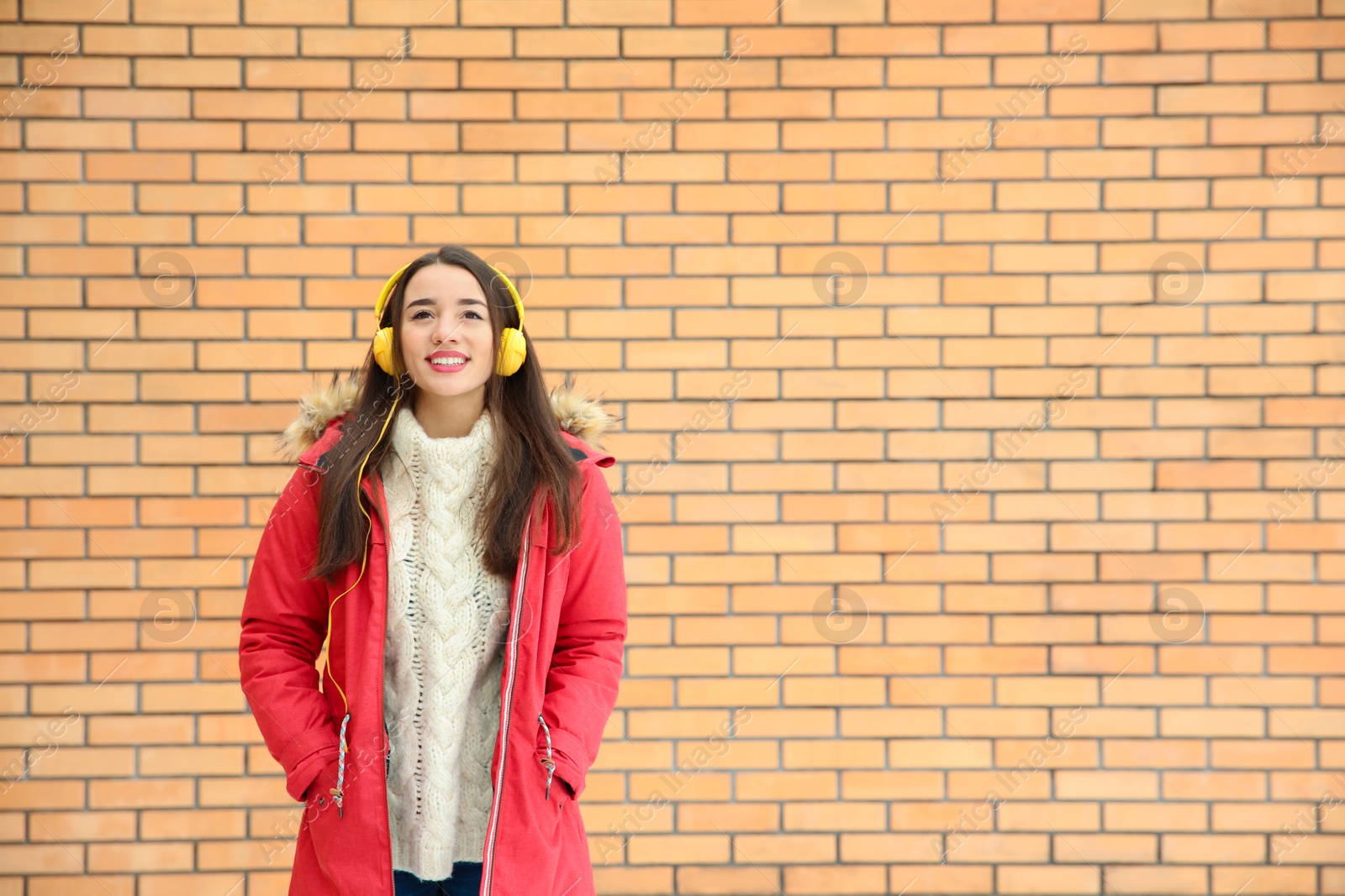 Photo of Beautiful young woman listening to music with headphones against brick wall. Space for text