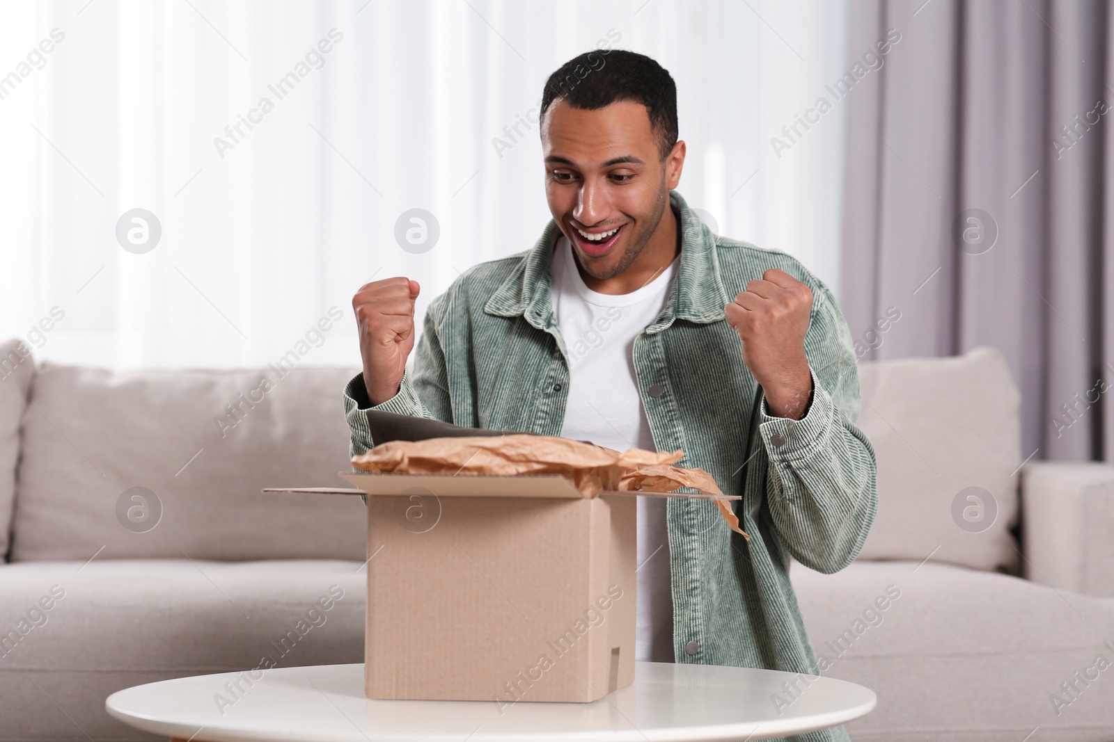 Photo of Emotional young man opening parcel at table indoors. Internet shopping